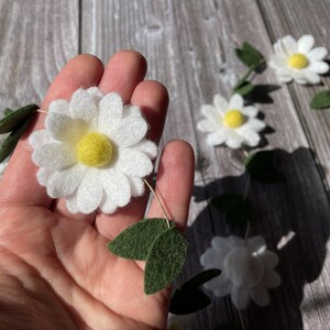 以下が含まれることがあります： 白いフェルトの花びらと黄色の中心部を持つデイジーガーランド、緑のフェルトの葉でアクセント。木製の背景に表示されています。デイジーの花の直径は約7.6cmです。