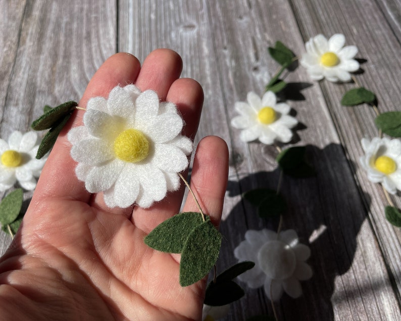 以下が含まれることがあります： 白いフェルトの花びらと黄色の中心部を持つデイジーガーランド、緑のフェルトの葉でアクセント。木製の背景に表示されています。デイジーの花の直径は約7.6cmです。