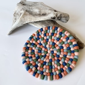 Op de afbeelding: Een ronde vilten onderzetter met een kleurrijk mozaïekpatroon. De onderzetter heeft een mix van kleine, ronde vilten ballen in roze, blauwe, groene en oranje tinten. De onderzetter ligt op een wit oppervlak met een stuk drijfhout op de achtergrond.
