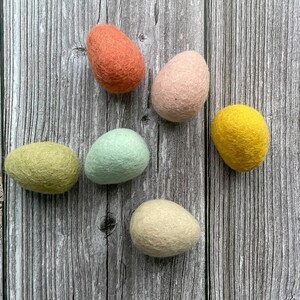 May include: Six colorful felt Easter eggs in shades of orange, pink, yellow, green, blue, and cream. The eggs are arranged on a weathered, gray wooden surface. These decorative eggs are perfect for Easter.