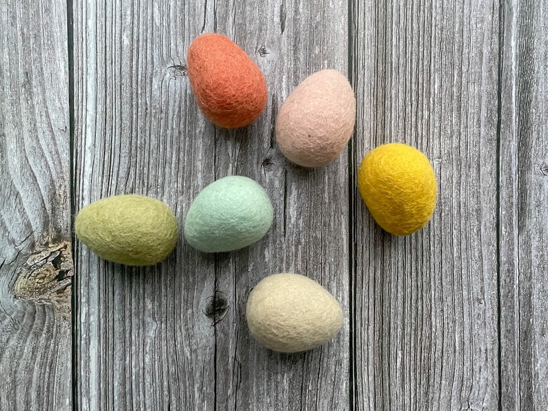 May include: Six colorful felt Easter eggs in shades of orange, pink, yellow, green, blue, and cream. The eggs are arranged on a weathered, gray wooden surface. These decorative eggs are perfect for Easter.