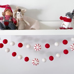 May include: A festive Christmas garland with red and white felt balls and peppermint candy accents. Decorative dolls and a snowman on a shelf above the garland. The garland is strung with white thread.