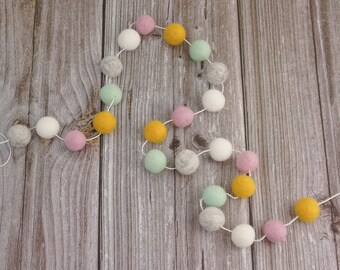 May include: A decorative garland of felt balls in pastel shades. The string of balls includes white, pink, yellow, grey, and mint green spheres, connected by white thread. The garland is shown against a wooden backdrop.