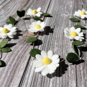 以下が含まれることがあります： 白いフェルトの花、黄色の中心、緑のフェルトの葉が付いたデイジーガーランド。 ガーランドは、春や夏の装飾に最適な、細い自然色の紐で繋がれています。 花は均等に配置されています。