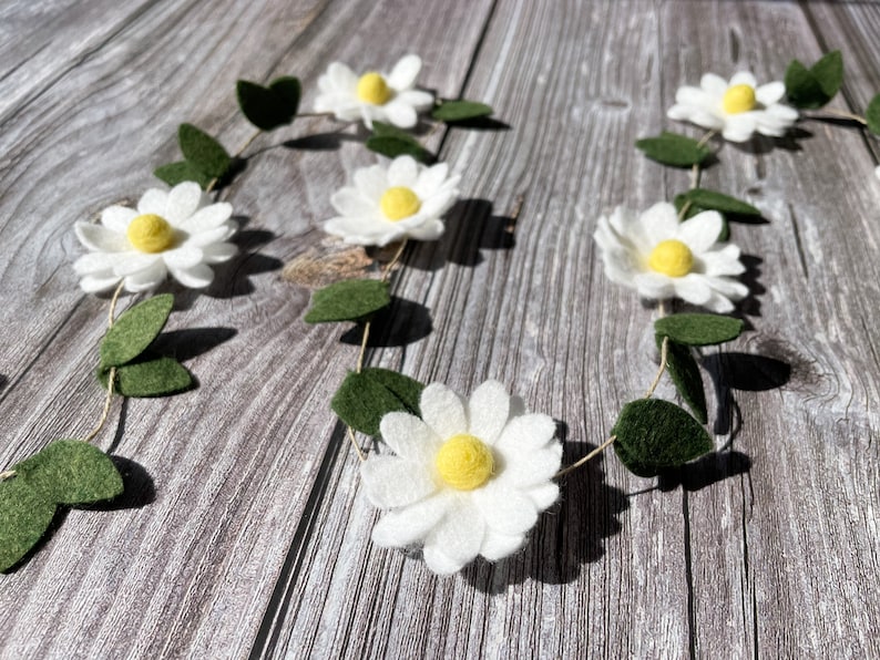 以下が含まれることがあります： 白いフェルトの花、黄色の中心、緑のフェルトの葉が付いたデイジーガーランド。 ガーランドは、春や夏の装飾に最適な、細い自然色の紐で繋がれています。 花は均等に配置されています。