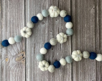 May include: A felt garland with white pumpkin shapes and felt balls in white, light blue, grey, and navy blue. The garland is displayed on a wooden surface, creating an autumnal decoration. The felt balls and pumpkins are of various sizes.