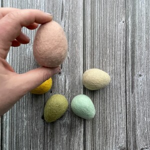 May include: A hand holding a light pink felt egg, with four other felt eggs in yellow, green, and light green colors. The eggs are on a wooden background.