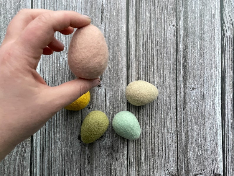 May include: A hand holding a light pink felt egg, with four other felt eggs in yellow, green, and light green colors. The eggs are on a wooden background.