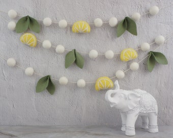 Puede incluir: Una guirnalda decorativa con bolas de fieltro blanco, hojas de fieltro verde y rodajas de limón de fieltro amarillo. La guirnalda está ensartada en un cordón fino de color natural. Una figura de elefante blanco está en primer plano.