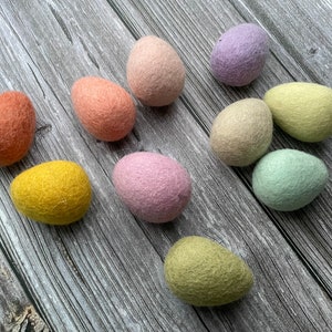 May include: A collection of ten colorful felt Easter eggs in shades of orange, pink, yellow, green, and purple. The eggs are arranged on a gray wooden surface, showcasing their soft, textured appearance. The eggs vary in size and shape.