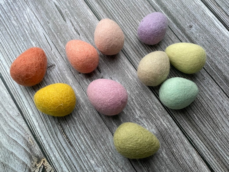 May include: A collection of ten colorful felt Easter eggs in shades of orange, pink, yellow, green, and purple. The eggs are arranged on a gray wooden surface, showcasing their soft, textured appearance. The eggs vary in size and shape.