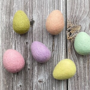 May include: Six colorful felt Easter eggs in shades of pink, green, yellow, and purple are arranged on a rustic wooden surface. The eggs have a soft, textured appearance, suggesting a handmade craft item.