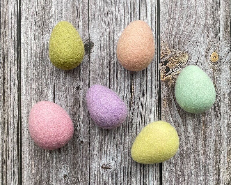 May include: Six colorful felt Easter eggs in shades of pink, green, yellow, and purple are arranged on a rustic wooden surface. The eggs have a soft, textured appearance, suggesting a handmade craft item.