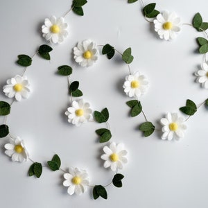 以下が含まれることがあります： 白いフェルトの花、黄色の中心、緑のフェルトの葉が付いたデイジーガーランド。 ガーランドは白い背景に対して流れるようなパターンで配置されています。 春または夏の装飾に最適です。