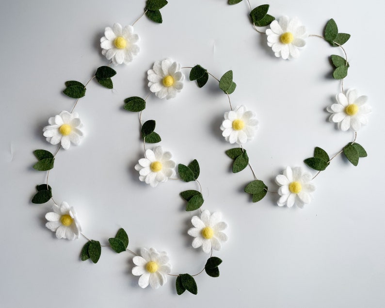 以下が含まれることがあります： 白いフェルトの花、黄色の中心、緑のフェルトの葉が付いたデイジーガーランド。 ガーランドは白い背景に対して流れるようなパターンで配置されています。 春または夏の装飾に最適です。