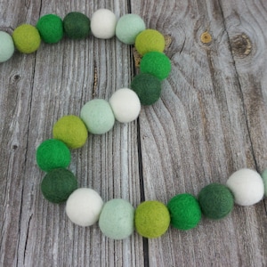 May include: A felt ball garland in shades of green and white, arranged in a curved pattern. The garland features various shades of green, including light green, dark green, and a muted green, interspersed with white felt balls. The garland is strung on a white string.