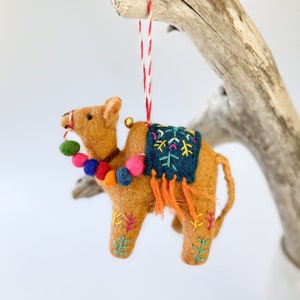 May include: A felt camel ornament in shades of brown, adorned with colorful pom-poms, embroidered details, and a dark blue saddle. The ornament hangs from a red and white string, with a small gold bell.