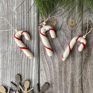 Puede incluir: Tres adornos de bast&oacute;n de caramelo blancos con rayas rojas y lazos para colgar. Cada bast&oacute;n tiene una peque&ntilde;a cuenta de madera en la parte superior. Los adornos est&aacute;n dispuestos sobre una superficie de madera con un copo de nieve y agujas de pino.