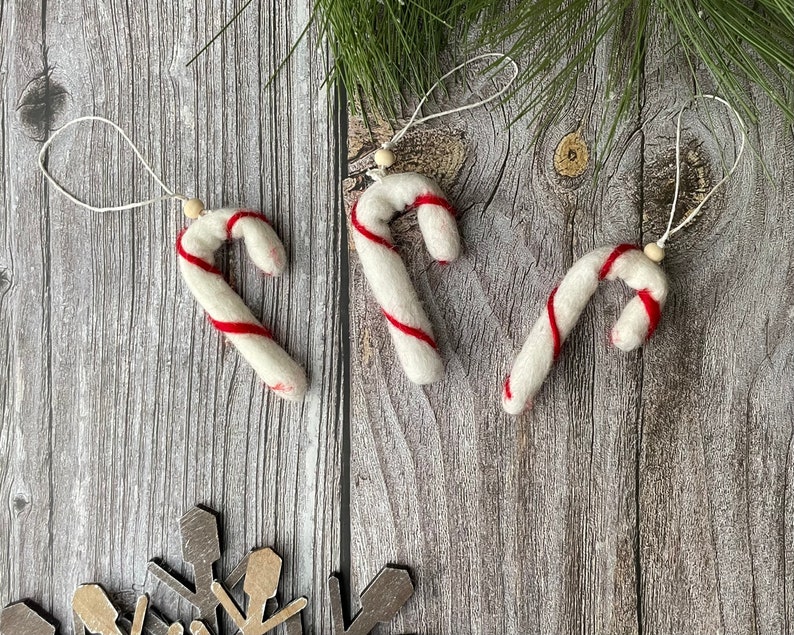 Puede incluir: Tres adornos de bast&oacute;n de caramelo blancos con rayas rojas y lazos para colgar. Cada bast&oacute;n tiene una peque&ntilde;a cuenta de madera en la parte superior. Los adornos est&aacute;n dispuestos sobre una superficie de madera con un copo de nieve y agujas de pino.