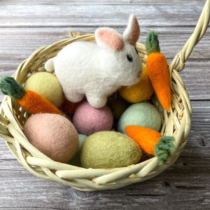 May include: A woven basket filled with colorful felt Easter eggs, two orange felt carrots, and a white felt bunny with pink ears. The basket has a handle and sits on a wooden surface.