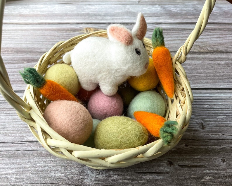 May include: A woven basket filled with colorful felt Easter eggs, two orange felt carrots, and a white felt bunny with pink ears. The basket has a handle and sits on a wooden surface.