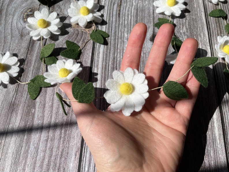 以下が含まれることがあります： 白いフェルトの花と黄色の中心、緑のフェルトの葉で作られたデイジーガーランド。ガーランドは細い自然色のコードに通されています。花は繰り返しのパターンで配置されています。