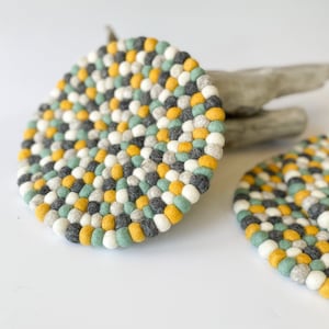 May include: Two round felt trivets with a colorful design. The trivets are made of small, tightly packed felt balls in shades of yellow, gray, white, and teal. The trivets are on a white surface with a piece of driftwood in the background.