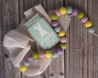 May include: An Easter decoration featuring a wooden sign with a bunny silhouette and the words "Hippity Hop" in white. A colourful felt ball garland in pastel shades of yellow, green, pink, and purple is draped over a burlap cloth, all set on a wooden surface.