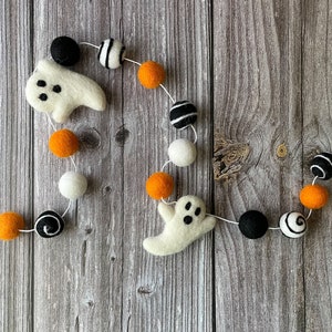 May include: A Halloween garland with felt ghosts, orange, black, white, and black and white striped balls strung on white thread. The garland is displayed on a wooden background.