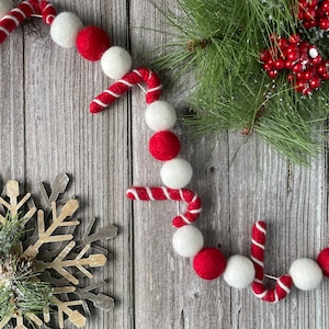 May include: A Christmas garland with red and white felt balls and candy cane decorations. The garland is displayed on a wooden surface, alongside a snowflake and evergreen branches with red berries, evoking a festive holiday atmosphere.