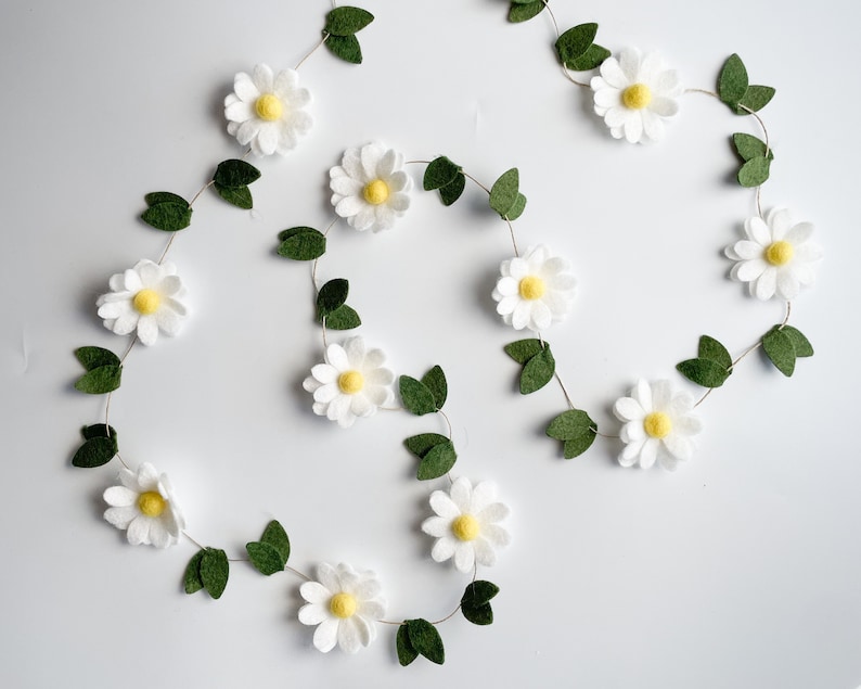 以下が含まれることがあります： 白いフェルトの花、黄色の中心、緑のフェルトの葉が付いたデイジーガーランド。 ガーランドは白い背景に対してループ状に配置され、陽気で春らしい美学を作り出しています。