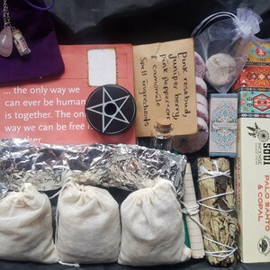 May include: A box filled with various items for a spell or ritual. The box includes a pentagram, herbs in small bags, a bottle of liquid, a small bag of white stones, a bundle of dried herbs, a box of incense, and a card with the text "Spell ingredients & camomile, pink peppercorn, Juniper berry, Pink rosebud."