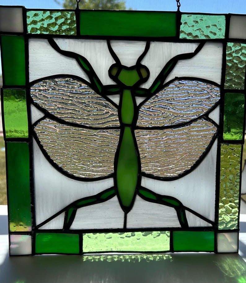 Pu&ograve; includere: Pannello in vetro colorato con un disegno di mantide religiosa verde. L'insetto &egrave; centrato su uno sfondo bianco, con ali argentate strutturate. Il pannello &egrave; incorniciato con vetro verde e trasparente.