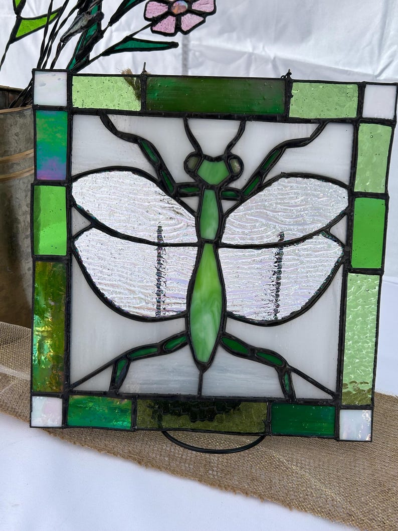 Pu&ograve; includere: Un pannello in vetro colorato con un disegno di libellula verde. La libellula ha un corpo verde e ali con un aspetto strutturato e iridescente. Il pannello &egrave; incorniciato con vetro verde e bianco, creando un pezzo decorativo.