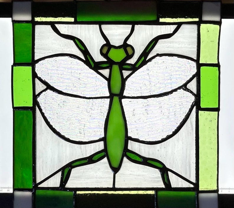 Pu&ograve; includere: Pannello in vetro colorato raffigurante una mantide religiosa verde con ali bianche, contornata di nero. L'insetto &egrave; centrato all'interno di un quadrato di vetro trasparente e verde, incorniciato da un bordo di vetro verde e nero.