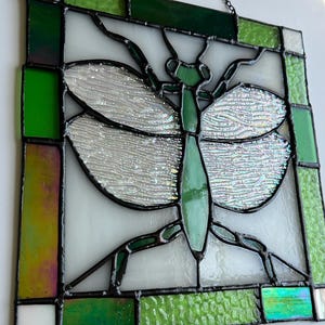 Pu&ograve; includere: Pannello in vetro colorato con un disegno di una mantide religiosa verde. L'insetto &egrave; centrato su uno sfondo bianco, con ali iridescenti e un corpo verde. Il pannello &egrave; incorniciato con vetro verde e trasparente e ha un bordo in metallo nero. Una catena &egrave; attaccata per appenderlo.