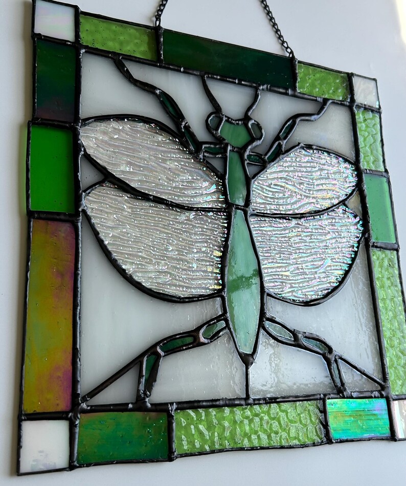 Pu&ograve; includere: Pannello in vetro colorato con un disegno di una mantide religiosa verde. L'insetto &egrave; centrato su uno sfondo bianco, con ali iridescenti e un corpo verde. Il pannello &egrave; incorniciato con vetro verde e trasparente e ha un bordo in metallo nero. Una catena &egrave; attaccata per appenderlo.