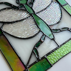 May include: Stained glass dragonfly art piece. The dragonfly features green body and head, with iridescent wings. The wings have a textured appearance. The piece is framed with black solder and includes green and white glass panels.