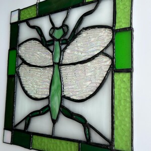 Pu&ograve; includere: Pannello in vetro colorato con un disegno di mantide religiosa verde. L'insetto ha ali iridescenti e un corpo verde, su sfondo bianco. Il pannello &egrave; incorniciato con vetro verde e bianco ed &egrave; sospeso da una catena.