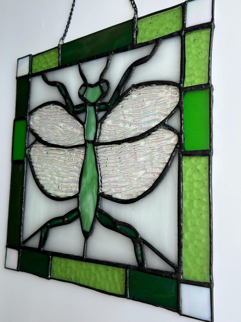 Pu&ograve; includere: Pannello in vetro colorato con un disegno di mantide religiosa verde. L'insetto ha ali iridescenti e un corpo verde, su sfondo bianco. Il pannello &egrave; incorniciato con vetro verde e bianco ed &egrave; sospeso da una catena.