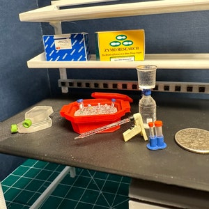 Könnte beinhalten: Ein Miniatur-Laborequipment mit einem roten Eiseimer, einer durchsichtigen Glasflasche mit blauem Verschluss, zwei blauen Reagenzgläsern mit roten Verschlüssen, einer weißen Plastikpipette, einem durchsichtigen Plastikbehälter und einer Silbermünze.