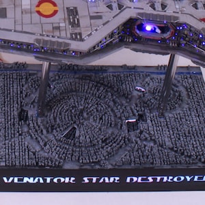 May include: A detailed model of a Venator-class Star Destroyer, a type of starship from the Star Wars universe. The model is displayed on a black base with a textured surface that resembles a starfield. The base also features the text "Venator Star Destroyer" in white lettering.