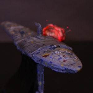 Pode incluir: Modelo detalhado de uma nave espacial, principalmente cinza com detalhes azuis, amarelos e vermelhos. A nave tem um design longo e elegante com uma seção frontal proeminente. Um efeito vermelho brilhante é visível no topo, sugerindo energia.