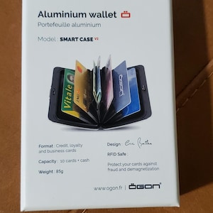 May include: A white box displays an aluminum wallet, model SMART CASE V2, with the text "Aluminium wallet" and "Portefeuille aluminium". The wallet holds credit, loyalty, and business cards. It weighs 85g and has a capacity of 10 cards plus cash.
