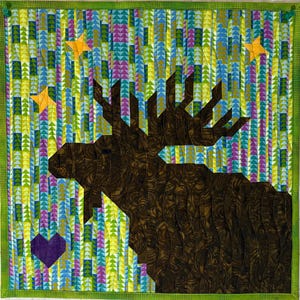 May include: A square quilt featuring a brown moose with large antlers against a colorful background. The background has vertical rows of triangles in blue, green, purple, and yellow. Orange stars and a purple heart are also included in the design. The quilt is framed with a green border.