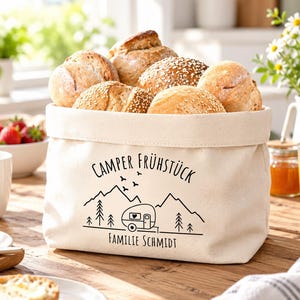 Könnte beinhalten: Ein cremefarbener Brotkorb aus Stoff, gefüllt mit verschiedenen runden Brötchen. Auf der Vorderseite befindet sich eine schwarze Strichzeichnung eines Wohnwagens, Bergen und Bäumen sowie der Text "CAMPER FRÜHSTÜCK" und "FAMILIE SCHMIDT".