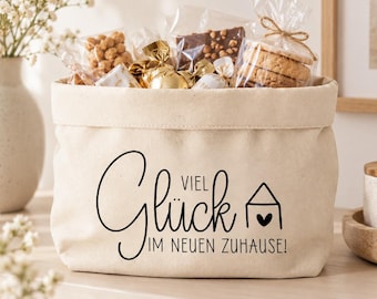 May include: A beige canvas storage basket filled with various snacks in clear plastic packaging. The front of the basket features black text that reads "Glück VIEL IM NEUEN ZUHAUSE!" along with a small line-art icon of a house with a heart.