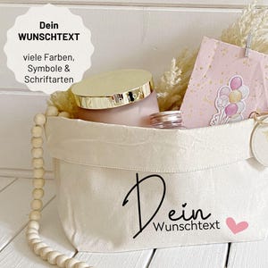 Könnte beinhalten: Ein weißer Canvas-Aufbewahrungskorb mit dem Text "Dein Wunschtext" und einem rosa Herz. Der Korb enthält ein rosa Glas mit goldenem Deckel, ein kleines Glas und eine rosa Geschenktüte mit Ballongrafiken. Eine Holzperlenkette hängt an der Seite.