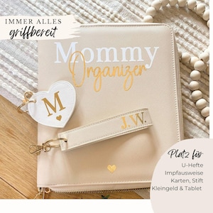 Könnte beinhalten: Ein beiger Mommy Organizer mit goldenen Buchstaben und einer passenden Handschlaufe. Ein herzförmiger Schlüsselanhänger mit dem Buchstaben "M" hängt am Organizer. Der Text "IMMER ALLES griffbereit" und "Platz für U-Hefte Impfausweise Karten, Stift Kleingeld & Tablet" sind ebenfalls sichtbar.