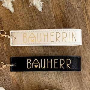Könnte beinhalten: Zwei Schlüsselanhänger mit goldener Beschriftung und einem Haussymbol. Der obere Schlüsselanhänger ist weiß mit dem Text "BAUHERIN". Der untere Schlüsselanhänger ist schwarz mit dem Text "BAUHEER".